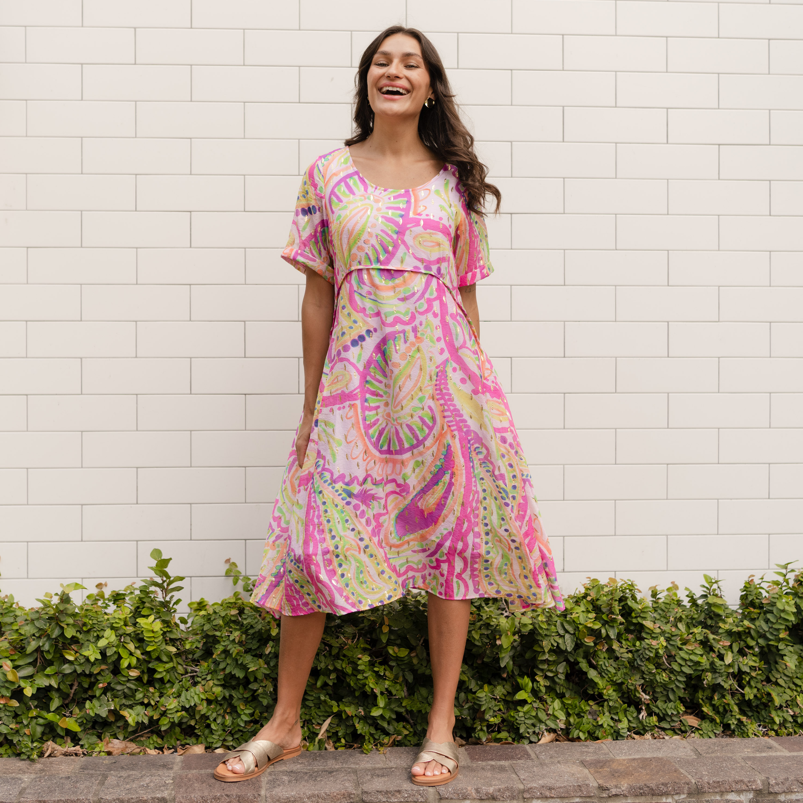 Brunette woman wearing a colourful A line dress in front of a white brick wall.