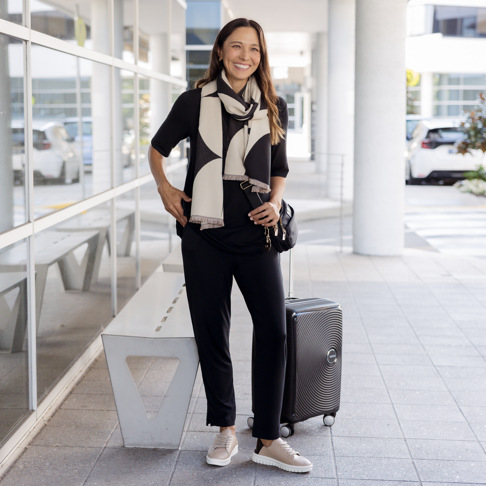 Brunette model wearing an airport outfit, consisting of a black top and pants, comfortable sneakers and a scarf. 