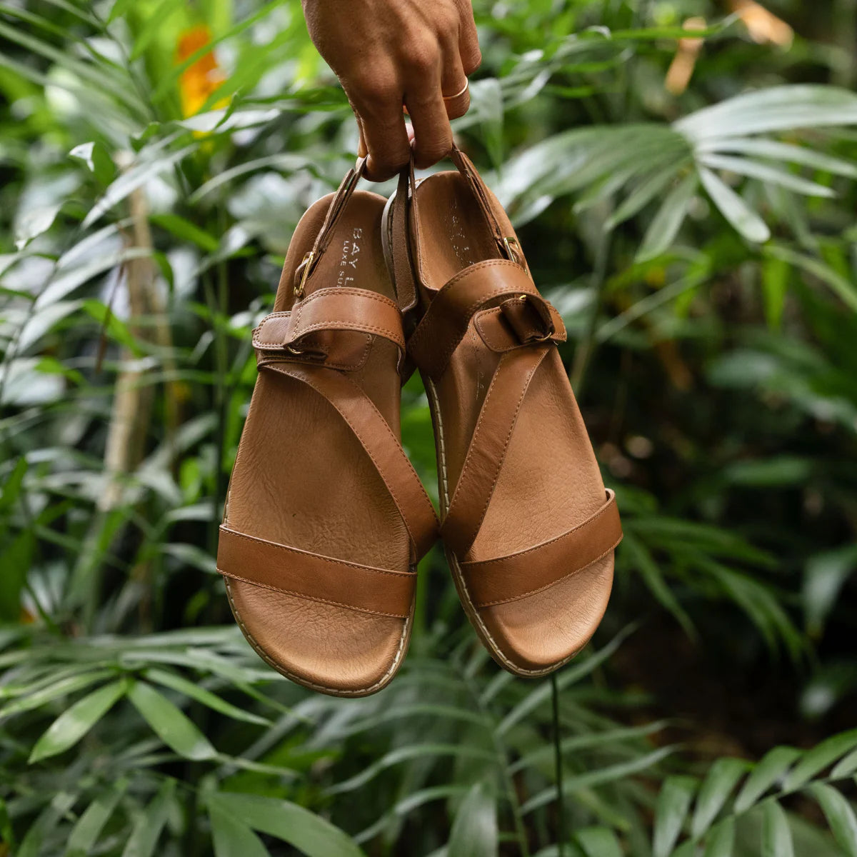 Woman holding tan strap sandals in front of a leafy garden.