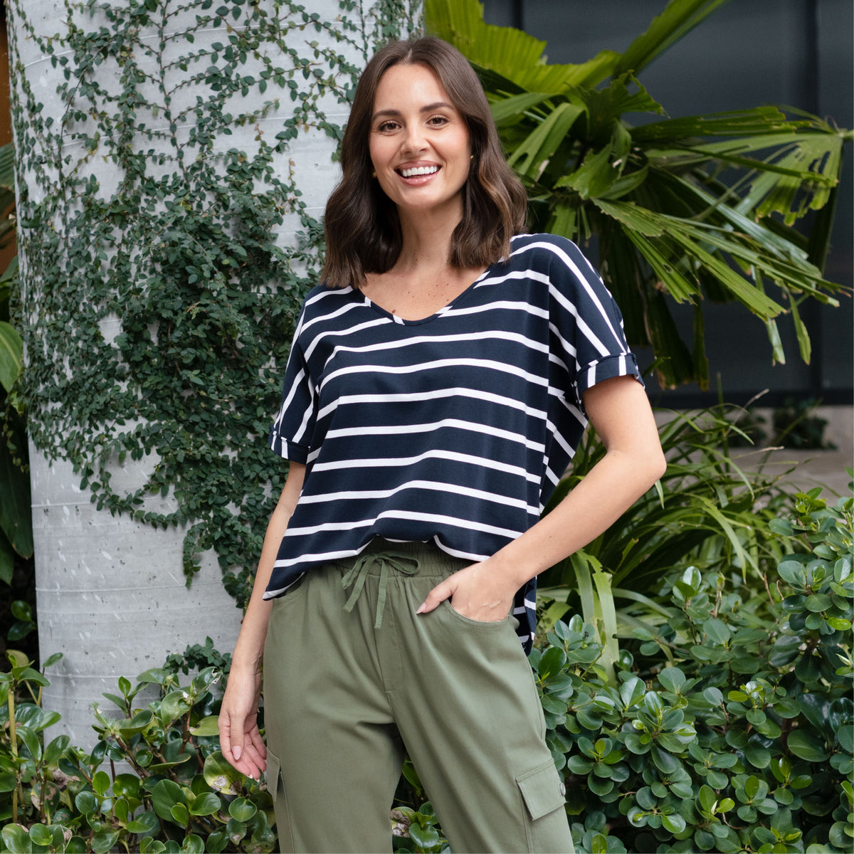 Brunette woman in loose striped oversized V-neck T-shirt smiles and poses with a hand in her pocket.
