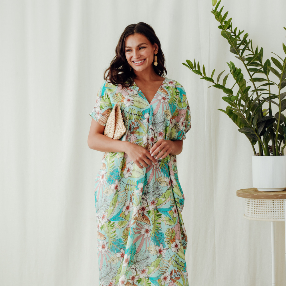 Brunette model wearing a green loral dress holding a clutch with a plant in the background