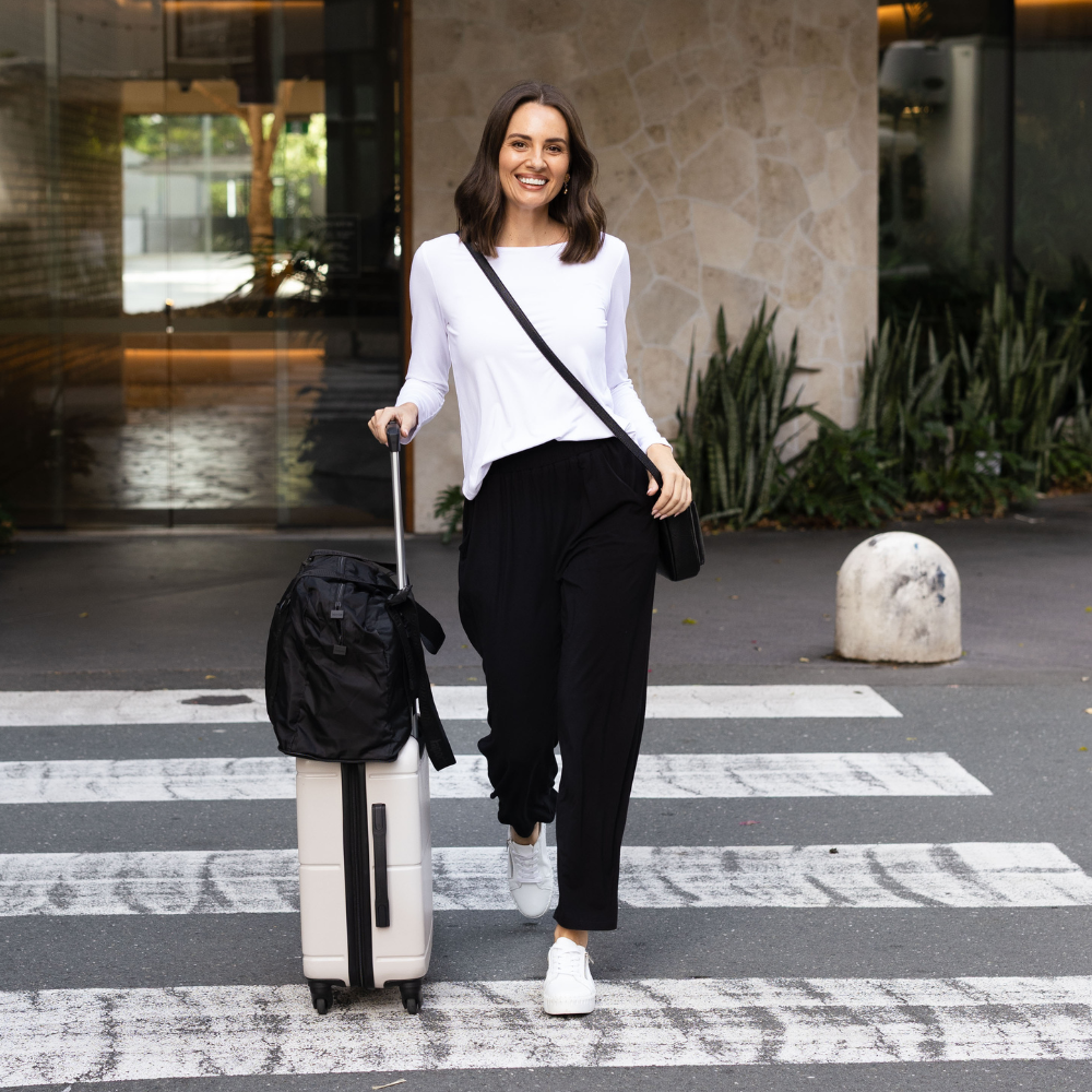 Brunette model wearing a long sleeve top and black pants wheeling a suitcase.