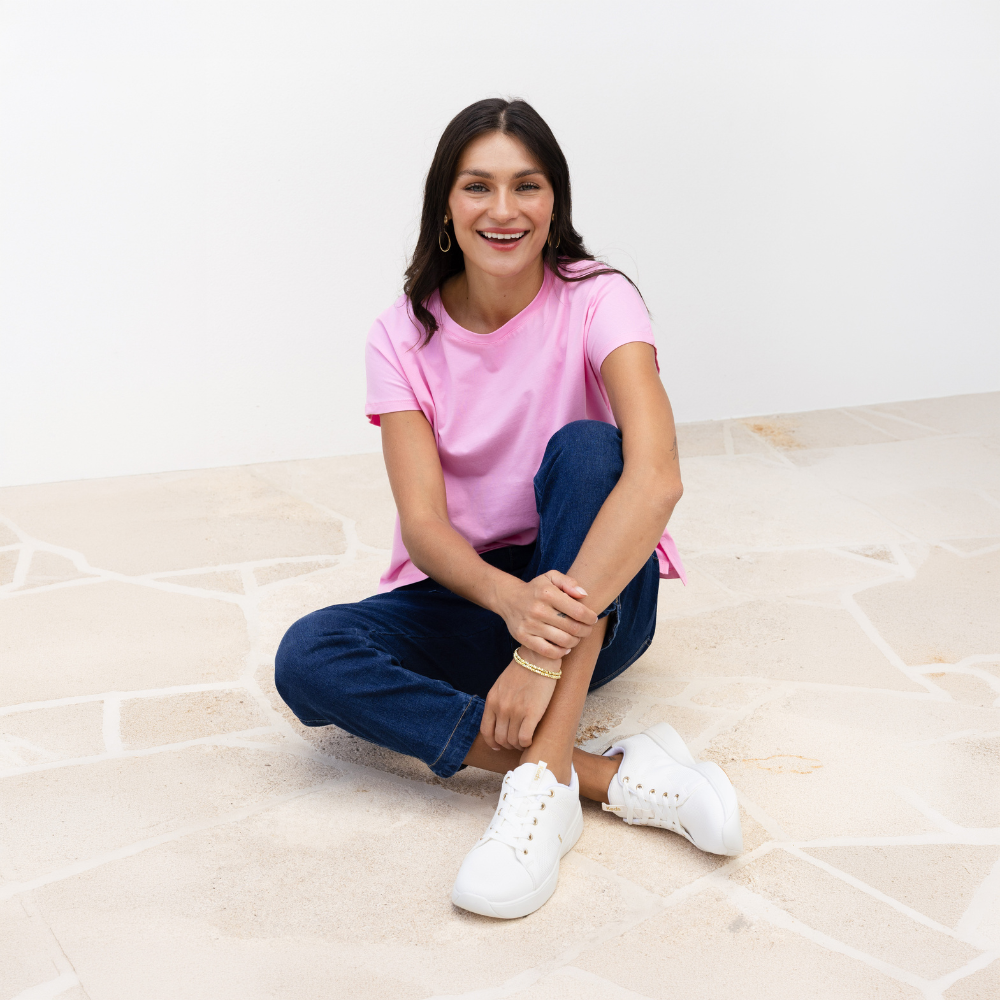A seated brunette model wearing a pink tee with blue jeans and white sneakers. 