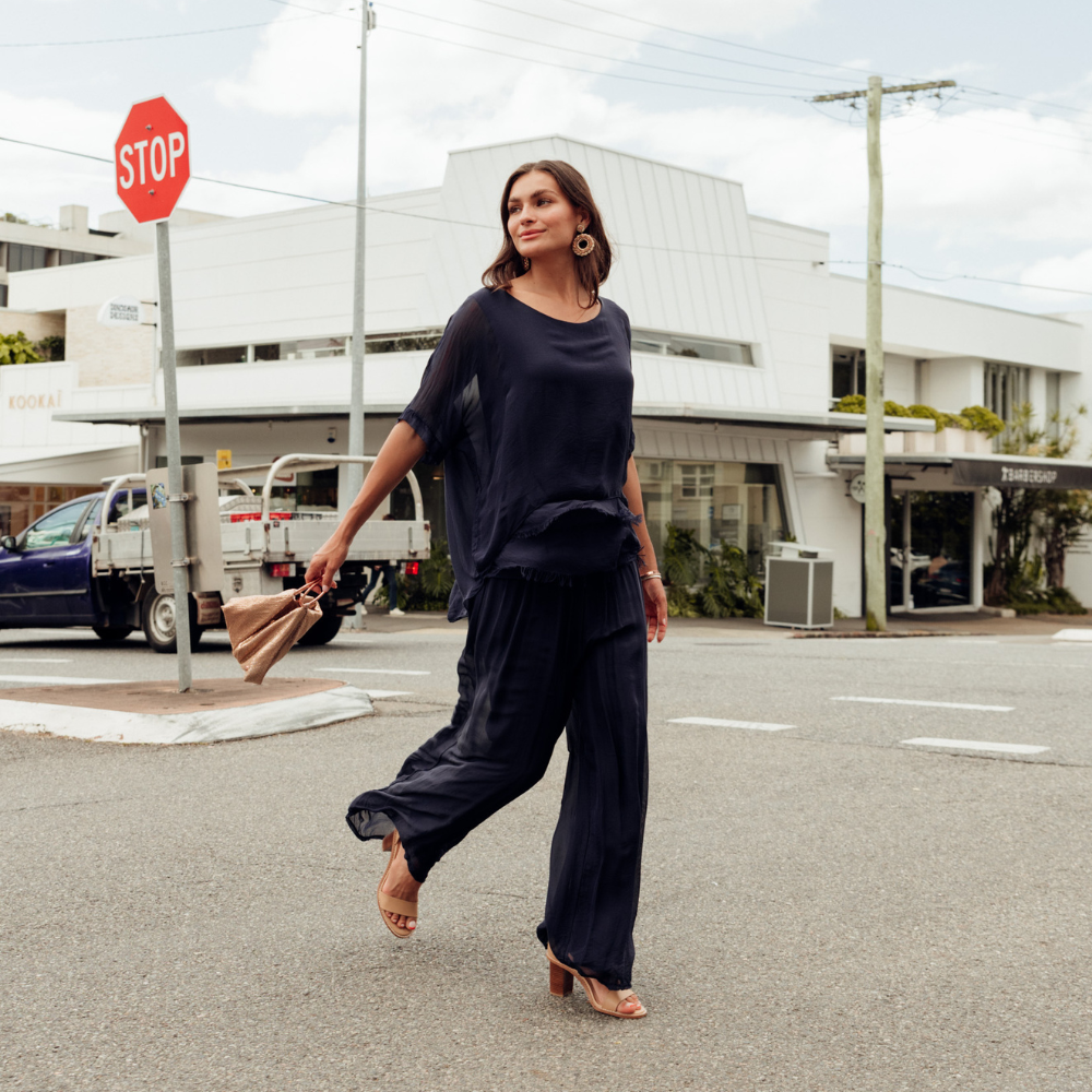 Brunette model wearing navy silk top and pants walking across the street holding a bag