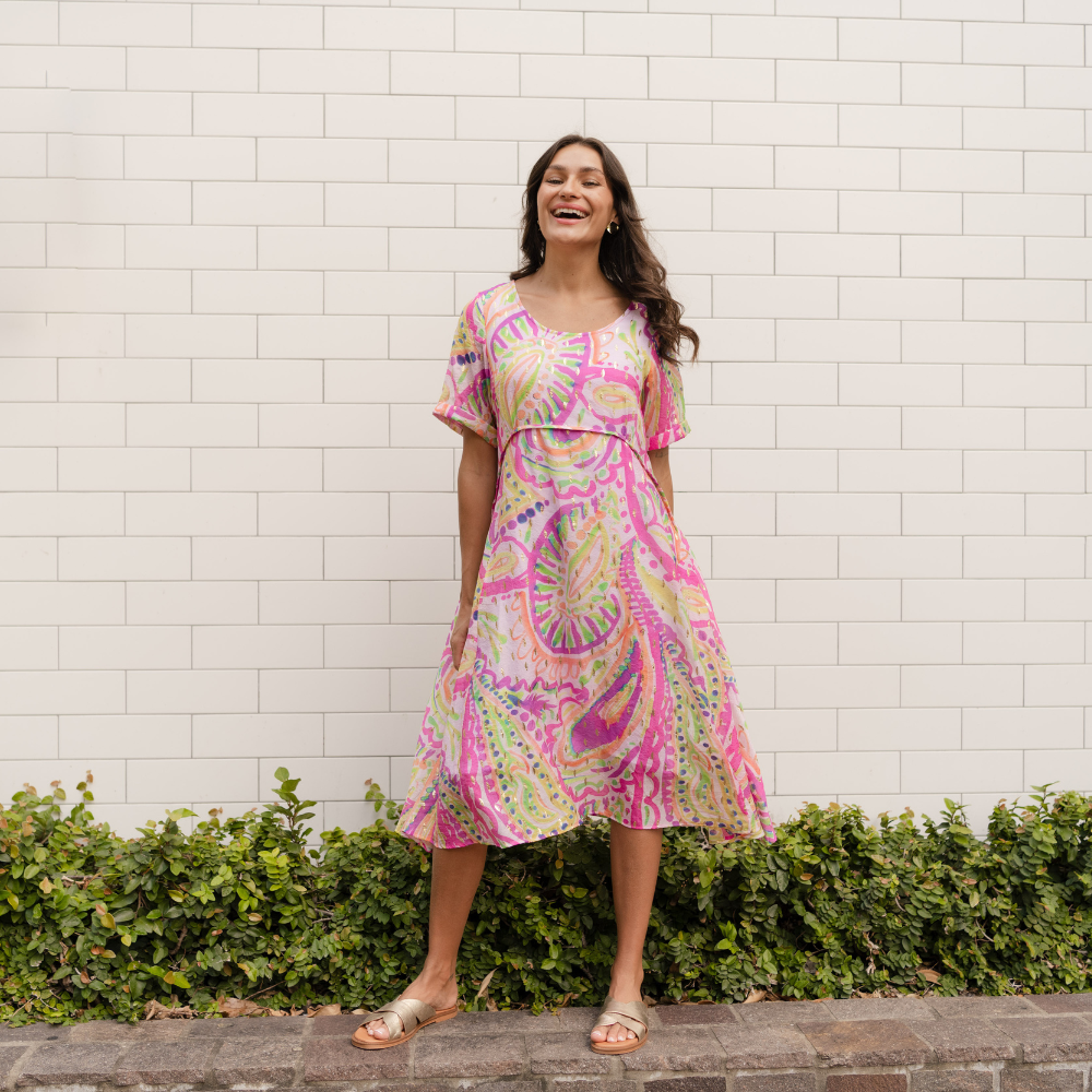 Brunette woman wearing a colourful summer dress in front of a white brick wall.