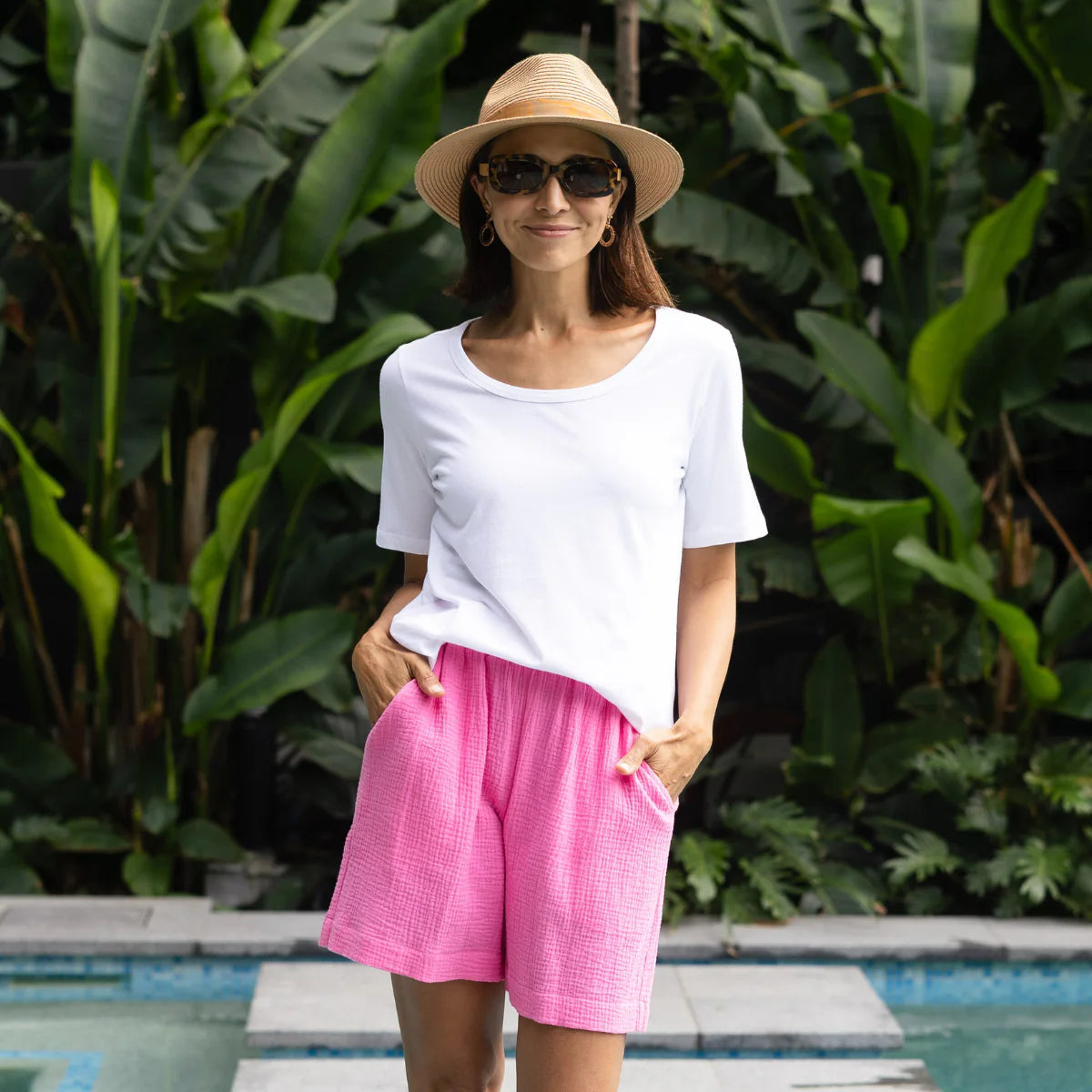 Brunette woman wearing a white tee, pink cheesecloth shorts, a woven hat and sunglasses on a tropical holiday.