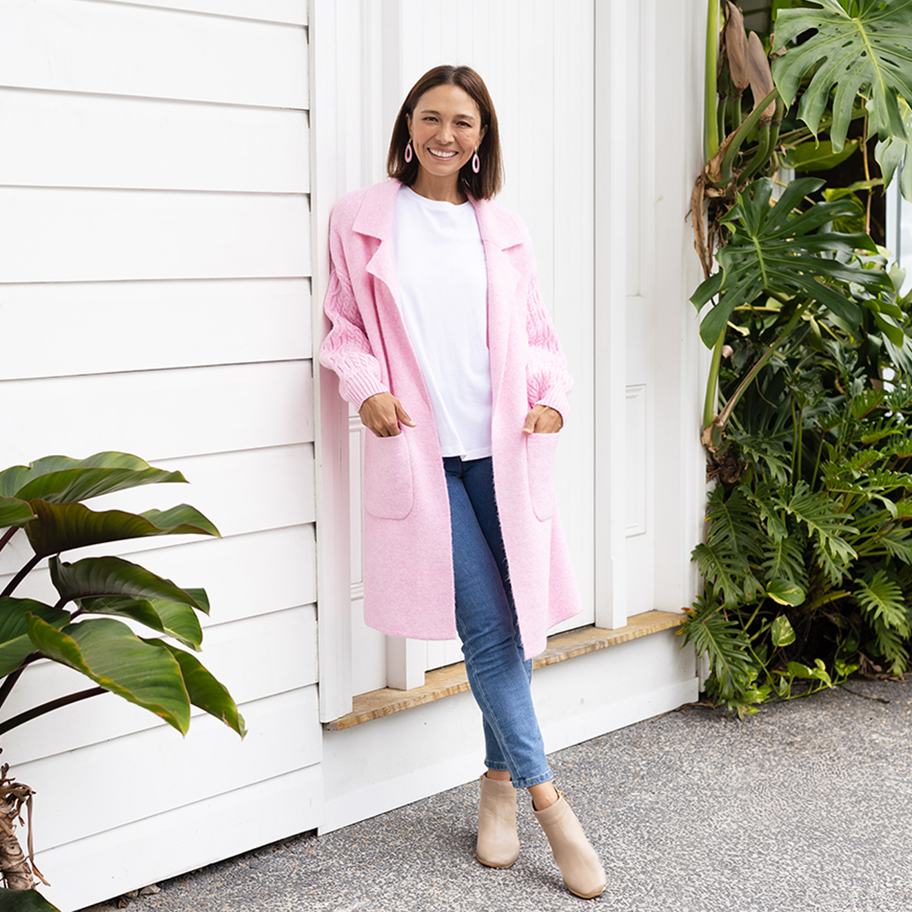 Brunette woman wearing a pink coatigan, blue jeans, and tan boots in front of a white house.