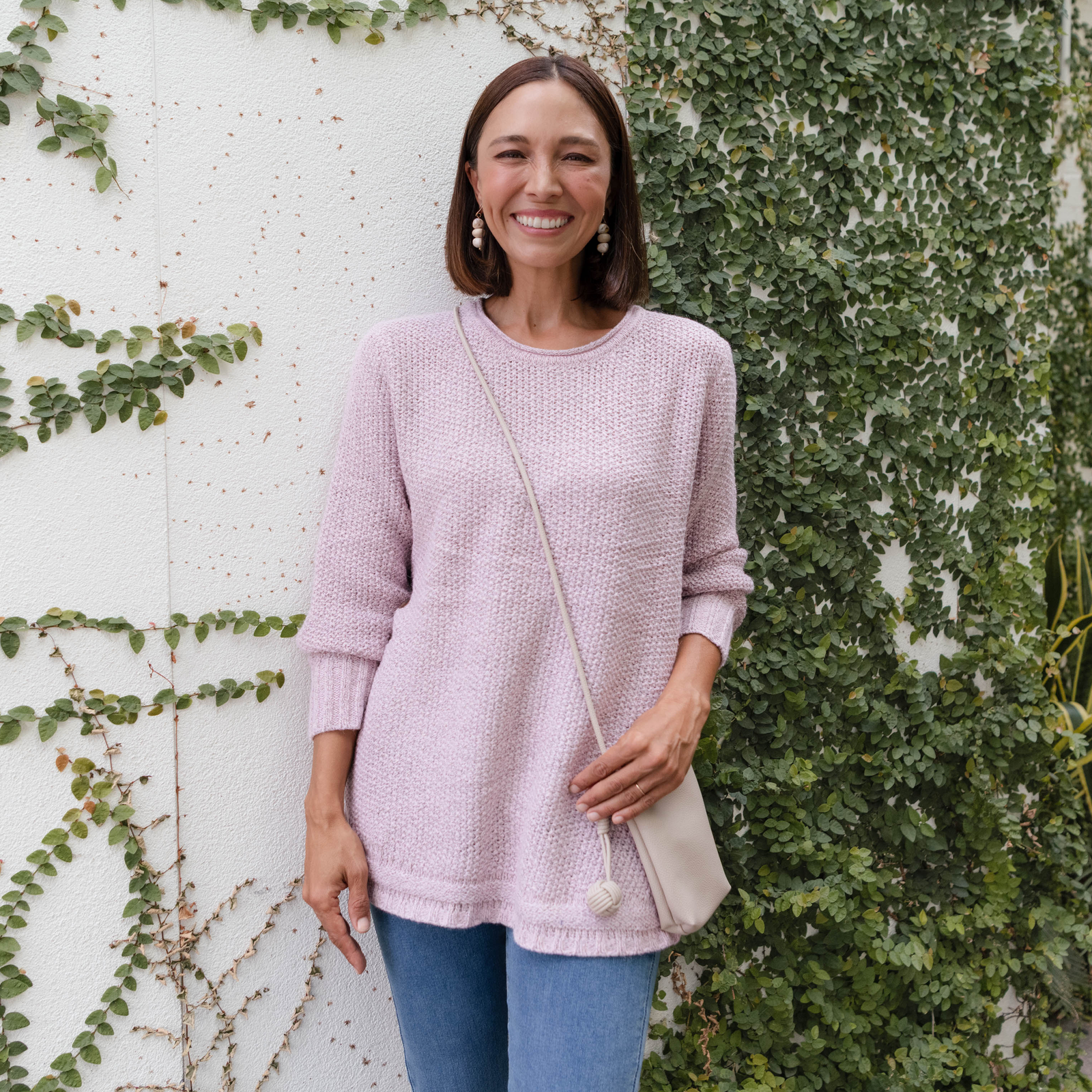 Brunette woman in a pale pink cardigan with a cream bag in front of a leafy wall.