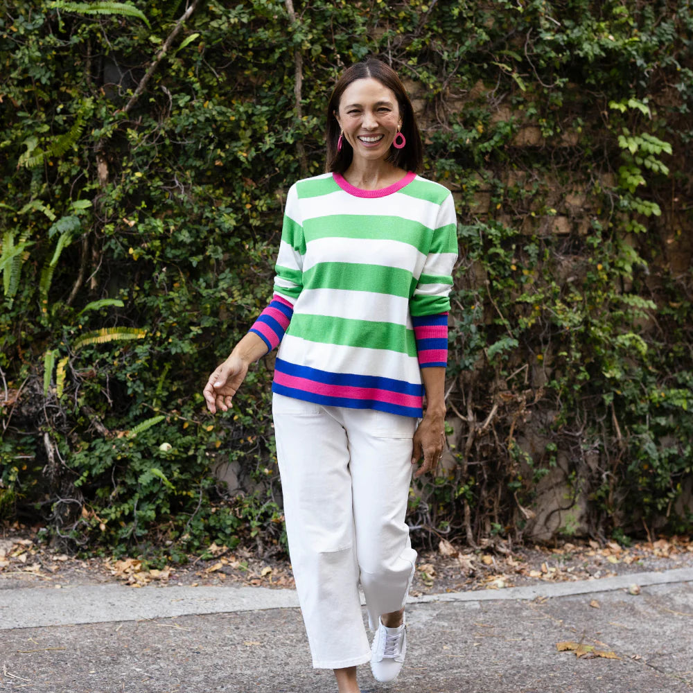 Brunette woman wearing a colourful green and white stripe knit jumper and white barrel jeans in front of a leafy wall.