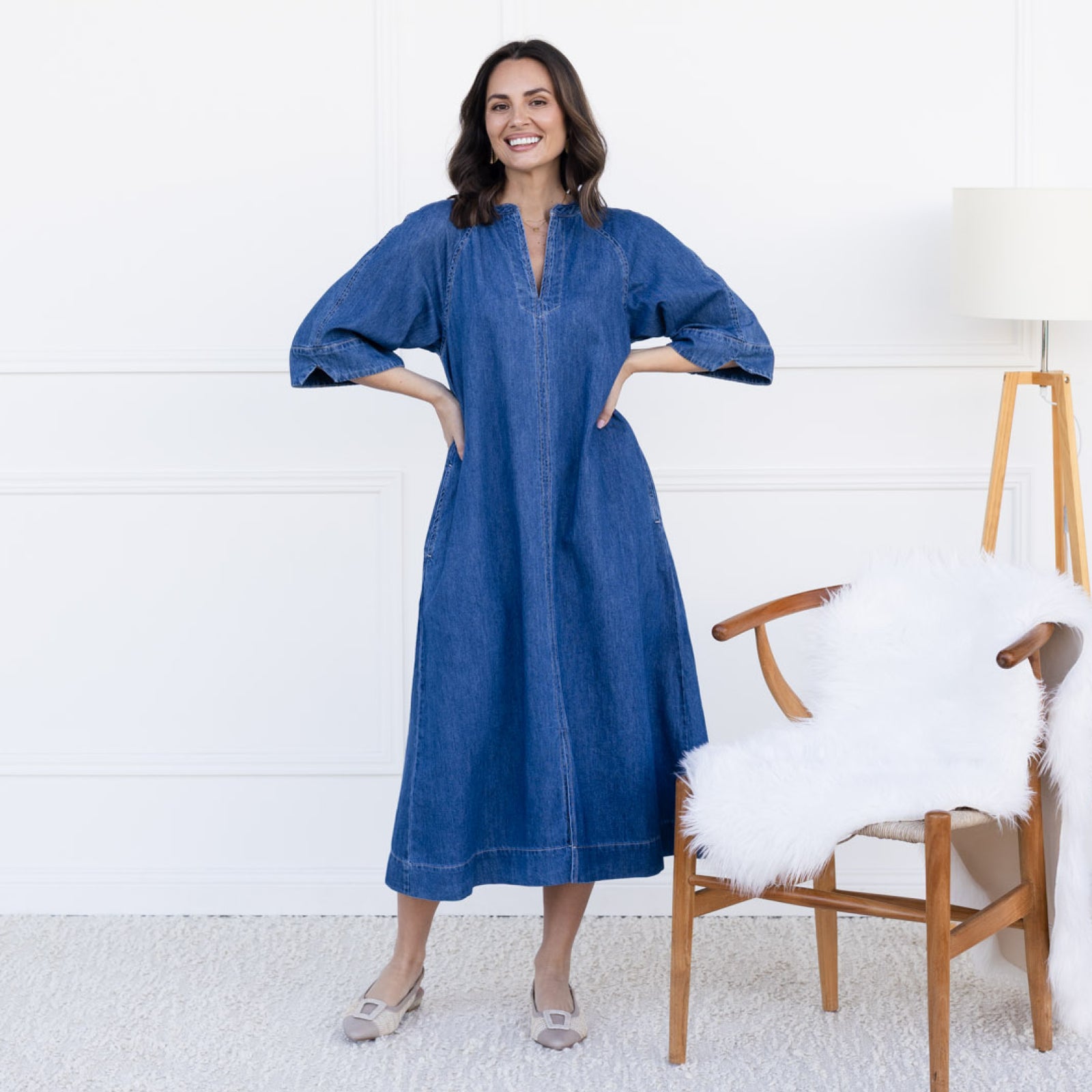 Smiling brunette woman stands in a denim maxi dress and flats.