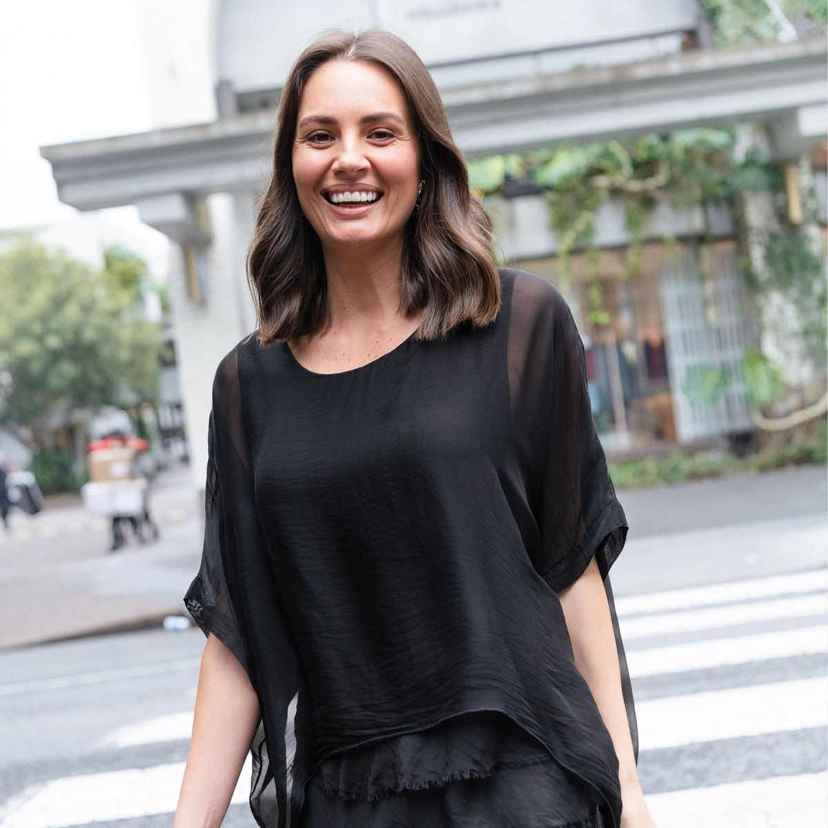 Brunette woman smiles crossing the road in a black wedding guest ensemble.