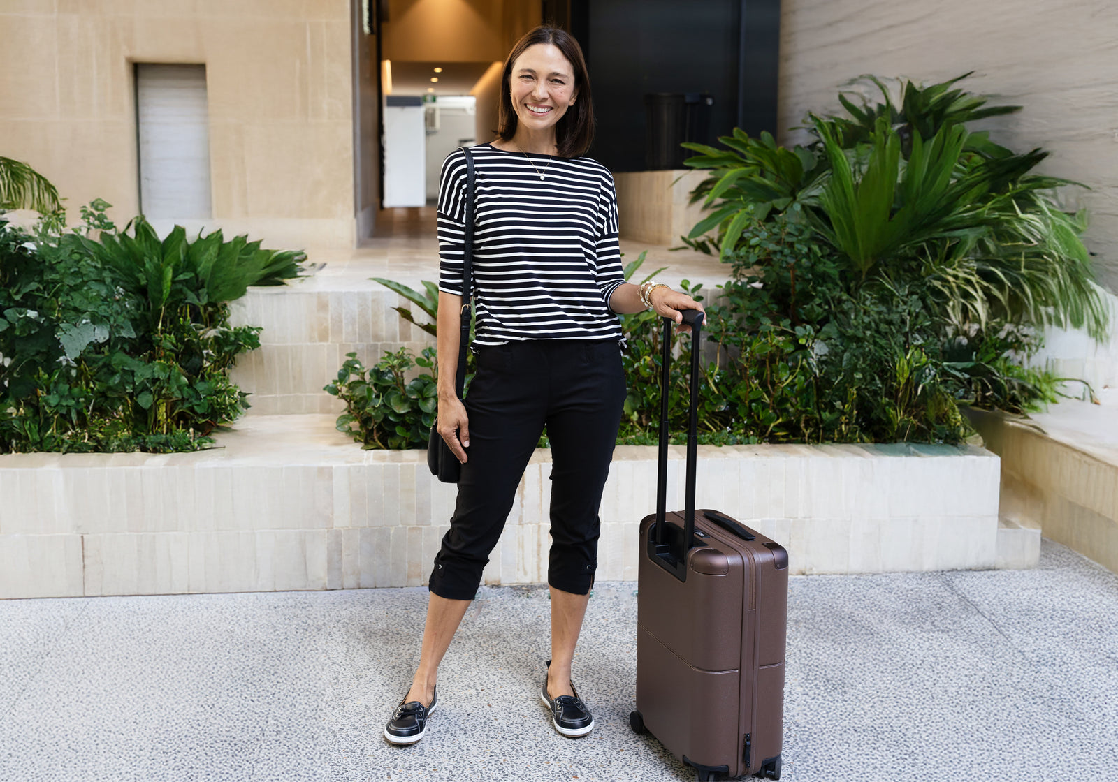 Brunette woman in a breathable monochrome travel outfit looks ready to go with a suitcase.