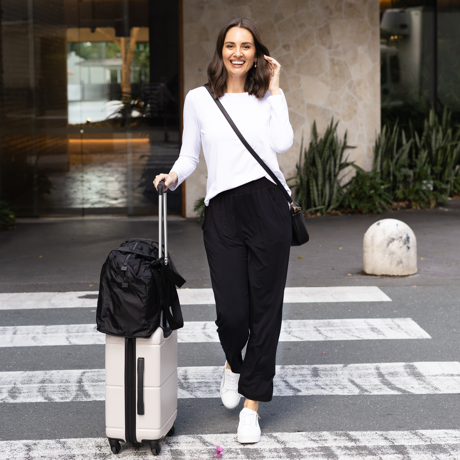 Brunette model wearing travel clothing, a long white sleeve tee and black pants, holding a suitcase.
