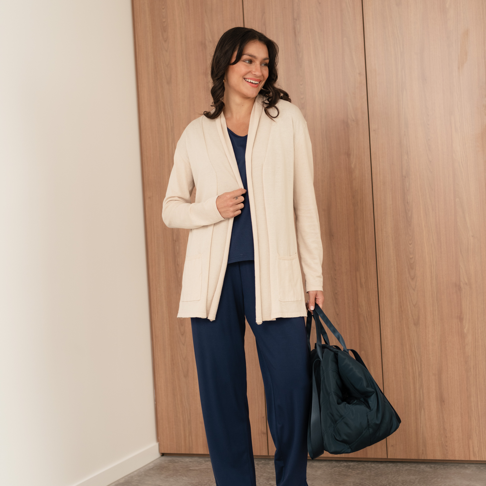 Brunette woman wearing navy travel pants and a beige cardigan holding a black duffle bag.