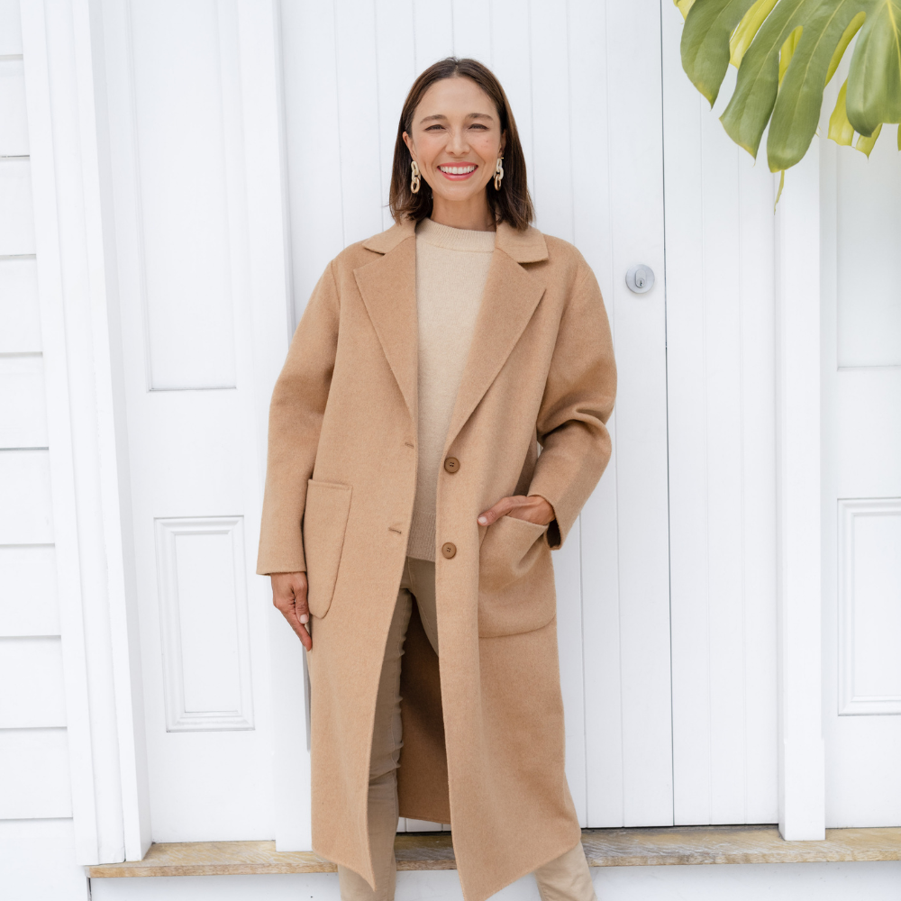 Brunette woman wearing a camel coloured winter coat in front of a white door.