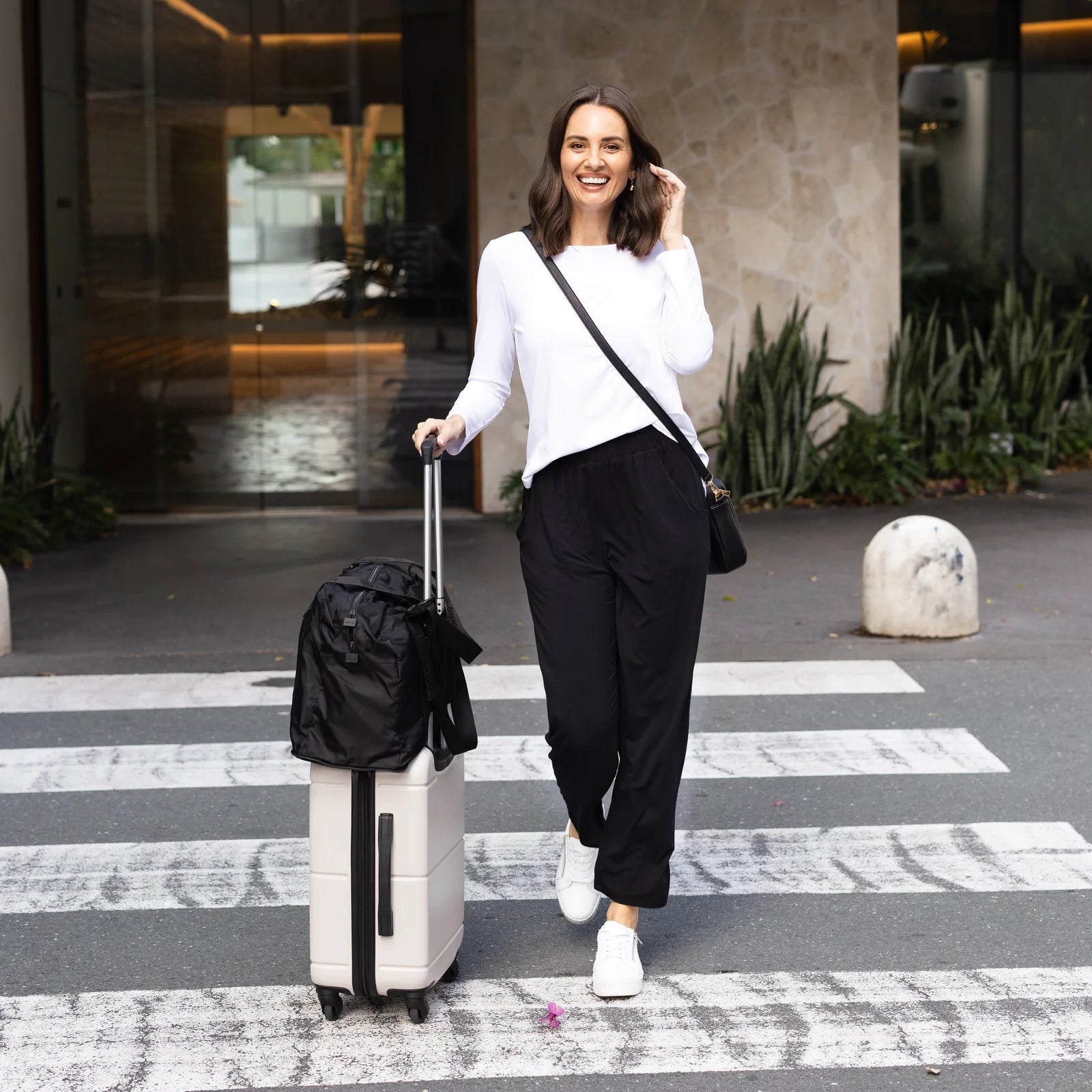 Brunette woman wearing a white top and black travel pants crossing the road with a suitcase.